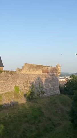 A wide shot captures a section of a stone fortress wall, bathed in the warm, soft light of early morning or late afternoon. The wall, constructed from large, rough-hewn stones, has patches of green ivy climbing its surface, suggesting age and the passage of time. The top of the wall is uneven, with several birds perched along its edge. To the right of the wall, a small, ornate tower with a domed roof rises, hinting at the architecture of a nearby town or castle complex. Beyond the wall and tower, a vista unfolds with trees and buildings visible in the distance, under a clear, pale blue sky. The ground in the foreground is covered in lush green grass, sloping downwards from the fortress. Several birds are also seen in flight against the sky. The overall impression is one of historical grandeur and natural beauty. The scene evokes a sense of peace and timelessness, enhanced by the golden light and the presence of the birds. The location is Dieppe, France.
