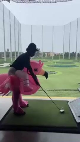 A person wearing an inflatable flamingo costume is at a Topgolf facility getting ready to swing a golf club. The person has on a black shirt, light-colored pants, and a black baseball cap. They are standing on a green rectangular platform, and a golf ball is on a tee in front of them. To the right of the person is a Topgolf touchscreen display. In the background is the golf driving range with the circular targets and netting surrounding the facility. The sky is overcast.
