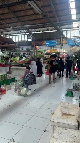 The video shows the interior of a busy open-air market. The market is mostly enclosed by a high metal roof with open sections. White tiles cover the floor. Several people are present, including a man in a plaid shirt who is kneeling down and packing vegetables into plastic bags. Other individuals are walking through the market, carrying bags of produce. Stalls are visible on both sides of the walkway, displaying a variety of fruits and vegetables. Blue signs with white lettering indicate the locations of facilities such as the "Mushola," "Posko Security," "ATM," and "Toilet." Green scales are placed near the produce. Some produce hangs from the stall roofs. The general atmosphere suggests a vibrant and bustling market scene.