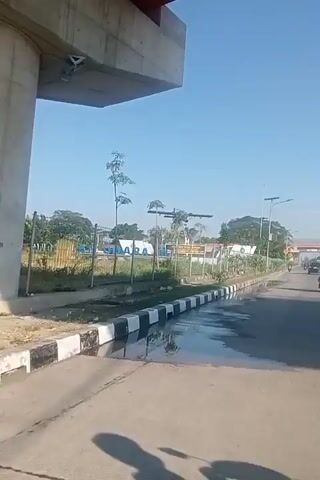 The video begins by showing a street with a shallow ditch on the left side, filled with standing water that reflects the clear blue sky. A fence runs along the side of the road, with trees and some buildings visible behind it. The camera appears to be moving, likely from the perspective of a vehicle. 

At 00:01, a drone is visible in the sky, positioned above the fence line. 

At 00:02, the camera continues to move forward, passing a solar-powered lamp post on the right side of the road. The ditch on the left remains visible.

At 00:03, a woman wearing a black headscarf and black clothing rides a black motorcycle towards the camera. She is passing the ditch and the fence.

At 00:04, the perspective shifts to a wider view of the road. Several motorcycles are seen traveling in the distance. The road appears to be a multi-lane highway or main road. Trees and some structures are visible on either side of the road. The sky is still clear and blue.

At 00:05, the video continues to show the road from the same perspective as 00:04. More motorcycles are visible in the distance, and the environment consists of trees and buildings along the side of the road. The overall scene is consistent with an outdoor, daytime setting on a sunny day.