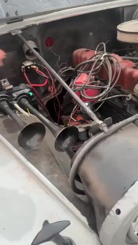 The video shows a close-up of the engine compartment of a grey Jeep. The hood is open, revealing a red engine with various wires and components. Two black horn-like objects are visible near the engine compartment. The camera pans out to show the front of the Jeep, including the grill, headlights, turn signals, bumper, and a portion of the tire. The environment appears to be outdoors, possibly in a residential area, as indicated by the pavement and grass in the background.