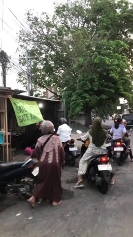 The video shows a street scene in Juwet Kenongo, Indonesia. It features several people riding motorcycles. The lighting suggests it is daytime, possibly late afternoon or early evening, as the light is somewhat diffused. The weather appears to be overcast or humid, common for tropical climates.

In the foreground, a person wearing a light brown hijab and a matching patterned garment is partially visible, facing away from the camera. They are on a motorcycle. To their right, another person in a patterned top and checkered pants, also on a motorcycle, is looking to their left.

Further into the scene, several other motorcycles are visible. One person in a white shirt and a patterned headscarf is riding a motorcycle. Another person, wearing a white shirt and blue shorts, is also on a motorcycle. The motorcycles are common models, suggesting everyday transportation.

The setting is a street with buildings and trees. On the left, there's a building with a green tarp extending from it, possibly a stall or shop. A utility pole with many wires is also prominent. The ground appears to be paved, with some dirt and debris. The overall atmosphere is that of a busy street in a populated area.

There are no discernible emotions expressed, as the focus is on the activity of people on motorcycles. The video captures a slice of daily life and movement within the town. No legible text is visible in the frame.