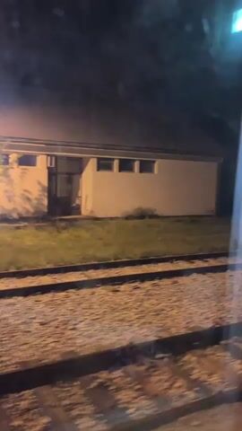 The video shows a view from a train window at night. The train is moving past a building illuminated by streetlights. The building is white with dark windows. A person in dark clothing is visible briefly near the building. The foreground shows the train tracks. The video ends with a close-up view of the scratched train window.