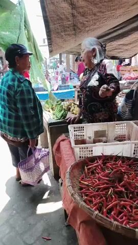 The video shows a bustling outdoor market scene. In the foreground, two women are conversing near a stall laden with fresh produce. One woman, wearing a green plaid shirt and a black hat, holds a purple woven basket. The other woman, with gray hair and wearing a patterned top, gestures with her hands as she speaks. Behind them, various fruits and vegetables are displayed, including bananas and a large basket of red chilies. The market appears to be under a makeshift roof, with various stalls and awnings visible. In the background, other shoppers and vendors are present, and motorcycles are parked nearby, suggesting a busy and active marketplace. The ground is dirt or gravel, with some debris scattered around. The overall atmosphere is lively and typical of an open-air market.