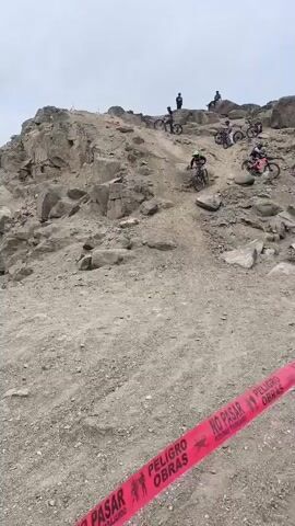 The video captures a downhill mountain biking event on a rugged, arid hillside overlooking the city of Trujillo, Peru, under an overcast sky.

The main subject is a mountain biker, wearing a full-face helmet, goggles, and protective gear, expertly descending a steep, dusty trail. The cyclist, riding a full-suspension mountain bike, navigates loose dirt and rocky terrain with precision. In the background, higher up the hill, several other cyclists and spectators are visible, suggesting a competition or organized riding session.

As the rider continues, the camera pans to reveal the extensive urban landscape of Trujillo stretching out below the hill. The trail passes by a concrete structure, possibly a ruined building, with a distinct blue-roofed building nearby. On one of the concrete walls, the text "RUTAS MEJES" is visible as graffiti. A person in a yellow shirt is briefly seen bending near this structure.

A section of the trail is marked with red and white caution tape, bearing the Spanish words "PELIGRO OBRAS" (Danger Works/Construction), indicating potential hazards or an active construction area. The biker performs a small jump or drop, kicking up a cloud of dust upon landing, demonstrating the dynamic nature of the sport. The scene effectively portrays the intense activity of downhill mountain biking in a challenging, semi-urban environment.