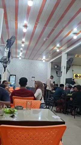 The video shows the interior of a busy restaurant with many people dining at tables. The ceiling is decorated with orange and white stripes, and the walls are white. There are fans mounted on the walls and lights on the ceiling. People are seated at tables, eating and talking. Some are wearing casual clothes, while others are wearing more formal attire. There are also several staff members circulating, serving food and drinks. The overall atmosphere appears to be lively and convivial.