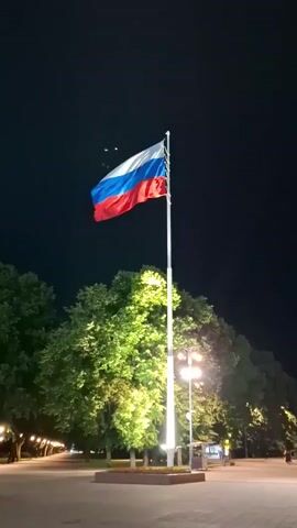 A Russian flag waves on a flagpole at night. The flag is illuminated and stands out against the dark sky. Trees are visible in the background, also lit by artificial lights. A few streetlights are also present. The overall scene is of a public space, possibly a park or square, at night with a prominent Russian flag.