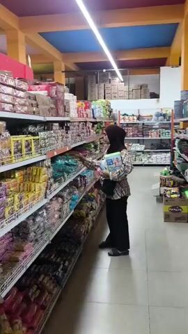 A person wearing a hijab and a plaid shirt is standing in a grocery store aisle, reaching for a product on the shelf. The aisle is filled with shelves of packaged goods, and the store appears to have high ceilings with exposed beams and lighting fixtures.