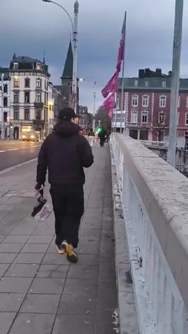 A person wearing a black hooded sweatshirt, black pants, and yellow sneakers is walking along the sidewalk of a bridge in an urban area. The person is carrying a rectangular object in their right hand. The bridge has a concrete railing on the right side. To the left, there is a street with cars and buildings. In the distance, there is a church steeple. The camera pans to the right, revealing a body of water with buildings on the opposite bank. The sky is overcast. There is a flag on the right side of the frame. There are other bridges visible over the body of water.
