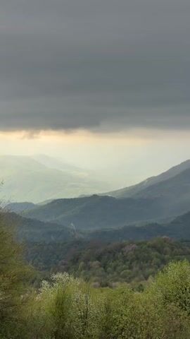 The video shows a mountain range under a dark, cloudy sky.  The mountains are covered in green trees.  A hazy light shines through the clouds, highlighting some of the mountain peaks.