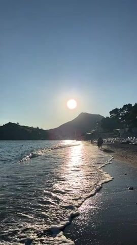 The video shows a beach scene at sunset. The sun is low in the sky, casting a warm glow on the water and sand. Waves are gently rolling onto the shore. In the background, there are hills covered with trees. A single person is walking along the beach.