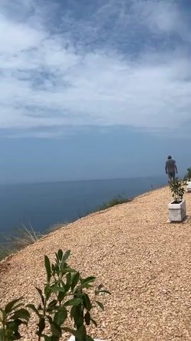 The video begins with a wide shot of a person walking away from the camera on a gravel path overlooking a vast, calm blue ocean under a partly cloudy sky. The path is lined with potted plants.

The camera then zooms in on a man wearing a dark t-shirt, glasses, and jeans. He turns to look at the camera and smiles slightly. He has tattoos visible on his arm.

Next, a woman with blonde hair and a white shirt, wearing a headscarf, smiles warmly at the camera. She is standing on the same path overlooking the ocean.

The camera then focuses on the woman's face as she laughs and turns her head away from the camera, gesturing with her hands. Another person's face, wearing sunglasses, is partially visible in the foreground, also looking towards the ocean.

The video concludes with a shot of the expansive ocean and sky from the cliffside. The weather appears to be sunny and warm. The overall mood is relaxed and enjoyable, suggesting a scenic outing. The location is identified as Dobra Voda, Montenegro. There is no visible text in the video.