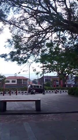 The video pans to the right, showing an outdoor scene with trees and buildings. In the foreground, there is a concrete bench. Behind the bench, there is a paved area with bollards and a motorcycle. In the background, there are several buildings, some with red accents, and a street with vehicles and more trees. The sky is cloudy. The camera then focuses on two tall, rectangular pillars with intricate patterns on them.