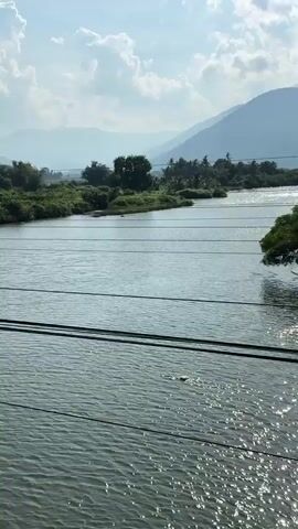 The video pans across a landscape. In the foreground, a river flows under a bridge, with sunlight reflecting off the water. A power pole with multiple wires crosses the frame. Beyond the river, there are lush green trees and fields. In the distance, mountains are visible under a bright blue sky with scattered white clouds. The camera moves to reveal rows of what appear to be train tracks, surrounded by fields and vegetation. The scene appears to be a rural area, possibly during the daytime.