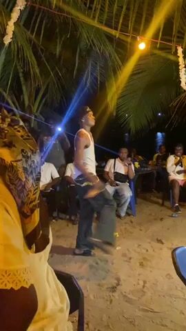 A nighttime beach party is in progress. A man in a white tank top, dark jeans, and sandals is dancing in the center, surrounded by people. Several are seated in chairs watching him. One person is filming the man with a smartphone. There are palm trees with lights strung between them, and the beach stretches out towards the water. Overall, the scene is lively and celebratory.
