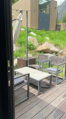 The video opens from inside a doorway, looking out onto a wooden deck. On the deck is a table with a marble-patterned top and a metal frame, accompanied by four chairs with metal frames and wooden slats, each with a grey cushion. To the left of the deck, a large, beige umbrella is partially visible. Behind the deck, there is a grassy slope with large rocks. Several modern, wooden cabins with glass elements are situated on the slope. String lights with illuminated bulbs are strung across the scene. The background features a dense forest leading up to a snow-capped mountain range under a cloudy sky. The camera pans slowly to the right, revealing more of the cabins, the forest, and the mountains.