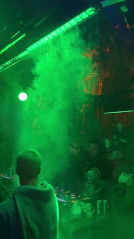 The video shows a crowded nightclub illuminated with purple lighting and hazy smoke. A DJ with short, light hair stands behind a mixing board, facing the audience. People are visible in the dimly lit crowd, some holding drinks and taking pictures. Decorative hanging elements are suspended from the ceiling, and a sign with white letters is visible in the background. The camera moves and focuses on the crowd, moving up towards the ceiling.