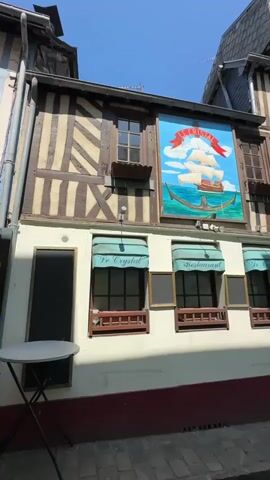The video shows the exterior of a historic building, likely a restaurant, on a sunny day. The building features half-timbered architecture with dark brown beams against a lighter colored plaster. There are several windows, some with green awnings that read "Le Crystal Restaurant". Above the windows, a large, colorful painting depicts a sailing ship with full sails, an anchor, and clouds against a bright blue sky. The painting is framed and mounted on the building's facade. To the left, a round white table with a black base and a black folding chair are visible on the cobblestone street. The overall impression is of a quaint and charming establishment.