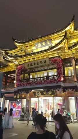 The video depicts a bustling street scene at night in what appears to be a commercial area with traditional Chinese architecture. The buildings are adorned with intricate designs and illuminated by golden and red lights. People can be seen walking around, some taking photos with their phones. There are shops and various commercial establishments lining the street. The sky is dark, indicating that it is nighttime. The scene evokes a vibrant and lively atmosphere.
