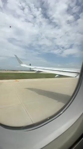 The video shows a view from an airplane window during takeoff. The airplane's wing is visible in the foreground, and the runway and surrounding landscape are visible in the background. The sky is cloudy. The airplane appears to be accelerating down the runway.