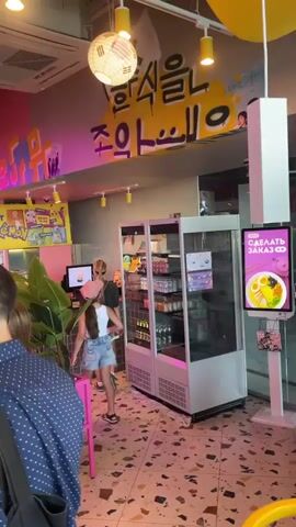 The video shows the interior of a brightly lit restaurant with vibrant pink and yellow decor. The walls are adorned with colorful graffiti and neon signs. Large green plants hang from the ceiling, adding to the lively atmosphere. In the foreground, a man with glasses wearing a blue and white patterned shirt is partially visible on the left. Behind him, another person is walking away. A woman with blonde hair, wearing a dark t-shirt and patterned shorts, stands near a refrigerated display case filled with drinks. In the background, customers are seated at pink tables, some interacting with each other and with staff. A large screen displays an image of a man in ornate clothing. The overall impression is of a modern, energetic dining establishment.