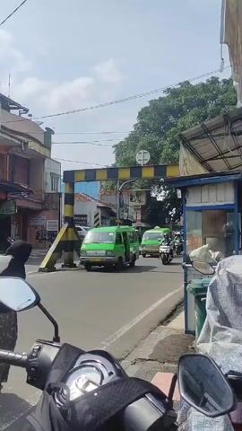 The video shows a street scene in a town or city. The footage appears to be shot from the perspective of someone sitting on a parked motorbike. In the foreground, we see the handlebars and mirrors of the motorbike.  The street is lined with buildings, some of which appear to be shops or small businesses. A black and yellow striped height restriction bar spans the road. Traffic, including green vans and motorcycles, is passing through. A woman is walking across the street carrying a large white sack on her back. She is wearing a dark top, a patterned skirt, and red sandals. The sky is partly cloudy. There is a small food stall on the side of the street with a blue frame and a corrugated roof.  Garbage bins are visible near the stall.