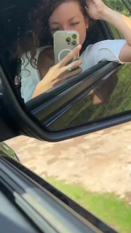 A woman with curly hair is seen in the reflection of a car's side mirror. She is wearing a white top and holding a smartphone, seemingly taking a selfie. Her arm is resting on the car door frame. The background reflects a dirt or gravel road and some greenery. The overall scene appears to be outdoors, possibly on a drive. The sound of wind can be heard.