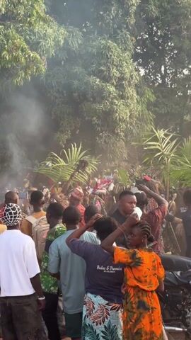 A large crowd of people is gathered outside in what appears to be a village. Most people are facing the center of the crowd, recording with phones. Many are wearing casual clothes, while others are dressed in colorful traditional attire, including elaborate headdresses.  There is smoke rising from the center of the gathering, obscuring the view somewhat. The environment is lush with green trees and foliage.  A few individuals are moving, but the overall impression is one of spectators watching an event.