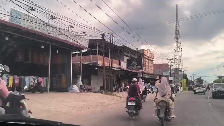 The video shows a view from inside a vehicle driving down a street in what appears to be a small town. Several people are riding motorbikes in the same direction as the vehicle.  Many of the women riding motorbikes are wearing hijabs. There are buildings with storefronts on either side of the street. Power lines crisscross overhead.  In the distance, there are cell phone towers. The sky is overcast with dark clouds. The traffic appears light.