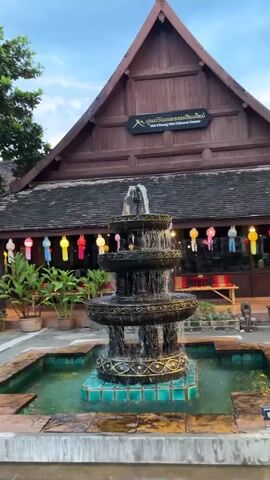 A tiered fountain in a courtyard is the central focus of the video. The fountain is made of dark stone and has water cascading down its levels into a turquoise pool. In the background, a traditional wooden building with a steep roof stands. Colorful paper lanterns are strung across the building's facade, suggesting an evening or festive setting. Green plants in pots are visible to the left of the building. The sky above appears overcast, indicating it might be dusk or a cloudy day. The video is shot from a static position, capturing the fountain and the building without any movement.