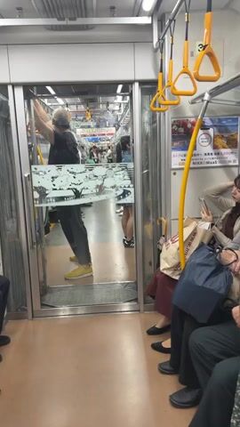 The video shows the interior of a crowded train car. The camera is positioned near a glass door, looking through it into the rest of the car. A man in a black shirt, dark pants, and yellow shoes is standing near the door, holding onto a handrail. He is wearing a beige baseball cap and a face mask. The door has a decorative white graphic of trees and flowers. Through the glass, many other passengers are visible, some standing and some seated. They are diverse in age and gender, and many are also wearing face masks. Handrails and straps are visible throughout the car, indicating it is a public transport vehicle. The overall impression is of a busy commute.
