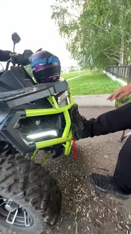 A person is shown placing their foot on the front of a quad bike. The quad bike has a lime green frame and black body. A helmet with purple and blue accents rests on the front of the quad bike. In the background, two young people are seated on a park bench. A fence and trees are also visible. The ground is covered with dirt and wood chips. The lighting suggests it is daytime.
