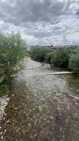 A short video shows a river flowing under a cloudy sky. The riverbed is visible through the clear water, filled with smooth, colorful stones. Trees line the banks of the river, and in the distance, houses with red roofs can be seen. A small waterfall is created by a man-made structure across the river. The video appears to be shot from a bridge looking downstream.