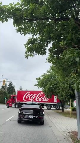 A black Kia EV6 is stopped at an intersection with a Coca-Cola semi-trailer truck behind it. The truck is red with white lettering and has images of Coca-Cola bottles on the back. The truck appears to be turning. The scene is on a street with trees and power lines. The sky is cloudy.