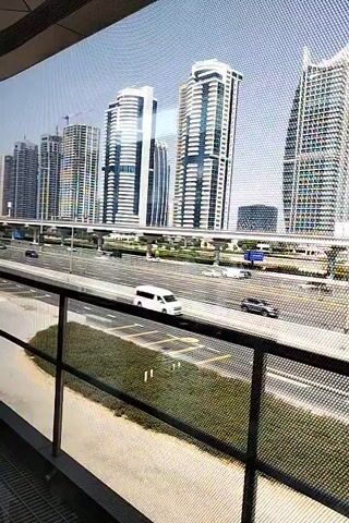 This is a video of a city skyline with tall buildings and a highway in the foreground. The camera is looking through a mesh screen, which creates a pixelated effect. There are cars driving on the highway, and some greenery around the highway. The sky is blue and clear, and the sun appears to be shining.