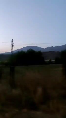 The video captures a landscape scene during twilight. The sky is a gradient of light blue and pale pink, suggesting either dawn or dusk. In the mid-ground, there are hills with a few trees. Two prominent electricity pylons stand against the backdrop of the hills. One pylon has a distinctive Y-shape at its top, while the other is a more standard lattice structure. The foreground is dominated by dry, brownish vegetation, which appears blurred due to motion, indicating the video was likely shot from a moving vehicle. The overall impression is of a rural or semi-rural environment transitioning from day to night. There are no people or animals visible in the footage.