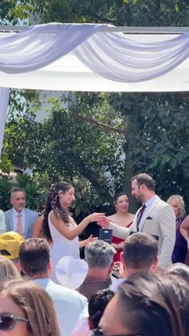 A wedding ceremony is taking place outdoors. A man in a light-colored suit and a woman in a white dress are standing under a white canopy. They appear to be taking shots of a clear liquid. The man raises his shot glass, and the woman does the same. They both drink their shots. There are guests seated in front of them, and more people standing behind them. Some of the guests are holding up their phones to record the event. The ceremony is surrounded by lush green trees.
