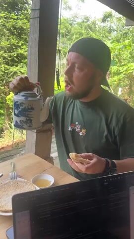 A man wearing a green T-shirt and a backwards baseball cap is sitting at a table outside, drinking directly from a blue and white ceramic teapot. He holds a piece of food in his other hand. There are plates of what appears to be food, a small dish of honey, and a laptop on the table. The background shows trees and foliage, suggesting an outdoor or semi-outdoor setting.