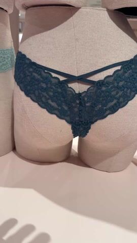 The video shows a display of women's lace underwear on mannequins in a store. A hand touches a pair of dark teal lace underwear on one of the mannequins. The underwear has a unique criss-cross design at the back. Another mannequin to the side displays light blue underwear with a lace trim. The mannequins are light beige and stand on a white surface. The lighting appears to be indoor store lighting.