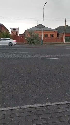 The video captures a street scene in Kyzylorda, Kazakhstan, likely during overcast daylight hours given the diffused lighting. The foreground is dominated by an asphalt road with visible white lane markings, bordered by a brick-patterned sidewalk.

In the background, residential buildings are visible. One prominent structure is a single-story house with an orange-toned brick facade and a dark, pitched roof. To its left, a smaller building with a red brick wall and a more utilitarian, boxy structure on top, possibly a shed or small outbuilding, is seen. Further to the right, another house with a green roof is partially visible. A streetlamp stands tall on the right side of the road.

The primary activity is traffic. A white sedan and a black SUV are shown driving on the road, crossing the frame from left to right. The motion blur indicates they are in transit. The scene appears to be a typical urban or suburban street. No people are directly visible in the frame, and no text is discernible. The overall impression is a quiet, functional street scene.