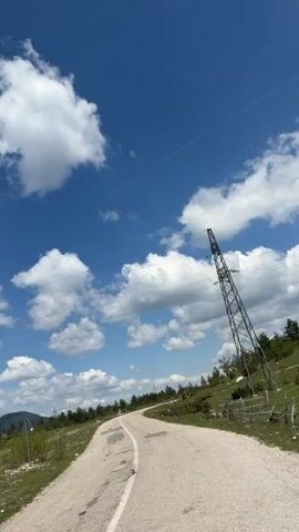 The video shows a view from a moving vehicle driving on a cracked asphalt road. The sky is blue with puffy white clouds. On the right side of the road, there's a grassy field dotted with shrubs and small trees, enclosed by a dilapidated wooden fence. An angled metal power line tower stands near the road, with power lines stretching across the sky. As the vehicle moves forward, the camera pans to the right, revealing a wider view of the green field, leading towards a distant mountain range under the cloudy sky. An abandoned building is seen further off, also with a power line nearby. The overall impression is of a rural or mountainous area with visible signs of infrastructure and a somewhat neglected landscape.