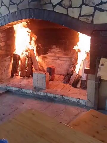 The video shows a close-up view of a large, stone-and-brick fireplace with a roaring fire. Several logs are burning, with bright orange and yellow flames reaching upwards. The fireplace is arched and made of rough-hewn stone and darker brick. The hearth is tiled with stone. In the foreground, a wooden table is partially visible, suggesting an indoor setting, possibly a restaurant or home. The fire appears to be actively burning and is the central focus of the video. There are no people or other animals visible in the frame. The lighting is warm and dominated by the light from the fire.