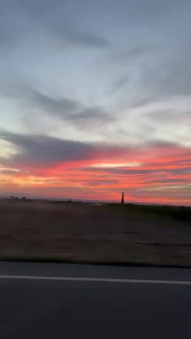 A video of a sunset over a rural landscape. The sky is filled with vibrant pink and orange clouds. In the foreground, there is a road with a white line. The landscape is blurred, suggesting motion, likely from a vehicle. There are fields and distant structures visible.