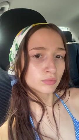 This is a close-up video of a young woman with long, dark hair and a bandana on her head. She is wearing a blue and white checkered bikini top and a necklace with a charm. She is in the backseat of a car, with car seats visible behind her. The woman is looking at the camera and making faces. The video is shaky, suggesting it was filmed on a phone.