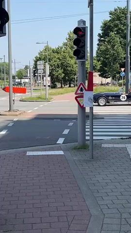 The media file shows a dark blue convertible sports car with white racing stripes driving through an intersection in Evere, Belgium. The car has the number "26" on its side. The time of day appears to be daytime, with a bright, sunny sky and clear weather. The setting is an urban environment with traffic lights, road markings, pedestrian crossings, and buildings in the background. Some visible text includes "DECATHLON" and "sky max" on buildings, and a directional sign pointing left with "INLEIDING" written on it. There is also text on the car's license plate, which appears to be "LO 4 M.955". The overall mood is one of casual observation of a classic car in a public space.