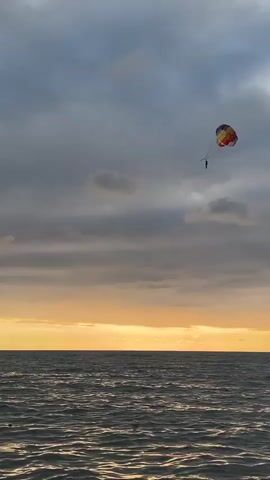 A person is parasailing over a choppy ocean at sunset. The sky is cloudy, and the sun is setting on the horizon, casting a warm glow over the scene. Several people are swimming in the ocean below. The parasail is multi-colored.