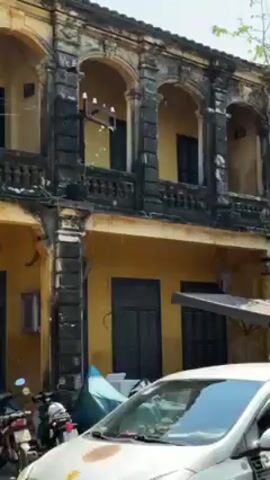 The camera pans past the facade of a yellow building in a tropical environment, revealing street vendors and businesses. The two-story building has dark green shutters on the windows and a decorative balcony on the upper level. The paint on the building appears weathered and faded. On the street, a white car is parked alongside scooters covered with tarps. A food cart stands near one of the buildings. As the camera continues to pan, a tree with hanging decorations and a red-and-white striped sign post with a yellow road sign appears. Further down the street is a small shop displaying items and a small snack shack. The day is sunny and bright.