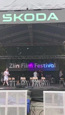 The video shows a stage with a banner that reads "Zlín Film Festival" and the logo of Škoda above it. A woman in a floral dress and black boots walks around the stage while a group of people stand near the back of the stage. The stage has a metal frame and a white tarp roof. There are chairs on the stage. A boy walks across the stage. Banners with the number "66" and the words "Zlín Film Festival" are attached to the sides of the stage frame. Speakers are also hanging from the stage frame. In the background, there are buildings and trees. The video appears to be taken from behind a fence.