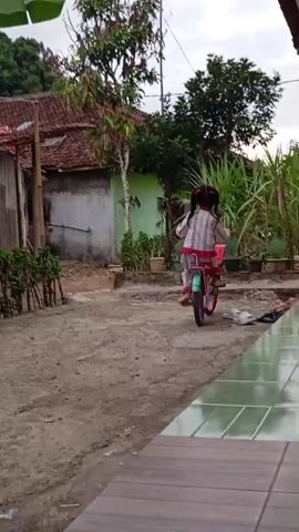 A child rides a bicycle on a dirt path in a village setting. The child is wearing a light-colored outfit with pink accents and has dark hair styled in pigtails. The bicycle is pink and blue with a basket on the handlebars. In the background, there are traditional houses with tiled roofs, lush green vegetation, and tall sugarcane plants. The ground is a mixture of dirt and gravel, with some scattered white objects and a patterned tile walkway visible in the foreground. The sky is overcast.