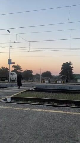 A person in a long black coat walks across a railway crossing as the sun sets. Overhead power lines are visible, along with a utility pole. In the background, trees and buildings can be seen. A white truck is parked on the left side of the frame.